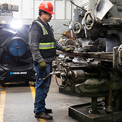Man in a safety vest working at a machine with a cool bass industrial fan blowing on him in the background